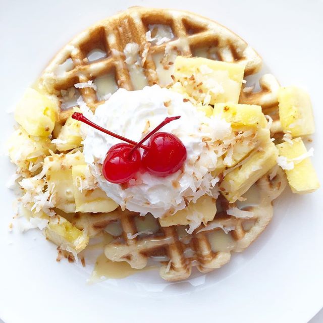 waffle on a white background topped with pineapple, whipped cream and cherries.